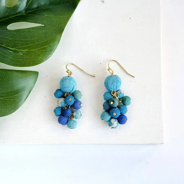 A photo of a pair of dangling earrings with blue sari kantha material wrapped beads in a cluster with a bigger blue bead on top made in India. 