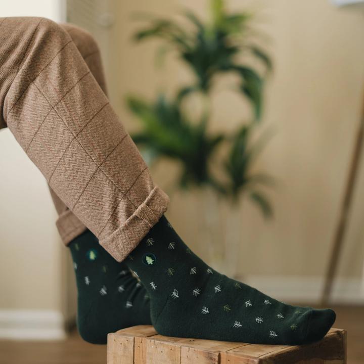 A photo of model wearing a pair of dark green socks featuring tiny trees in grey and green and a tree embroidery that help plant trees through non-profit donations with each purchase of these fair trade socks made in India.