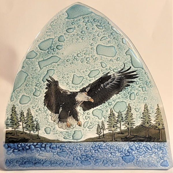 A photo of a nightlight made of recycled glass in Ecuador. This night light features a eagle flying with a background filled with light blue sky, green trees, and a deep blue water. 