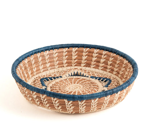 A photo of the Florinda basket, which is a handwoven basket made in Guatemala out of Pine Needles stitched with raffia in both natural and blue shades to create a central six-pointed star motif at the base of the Florinda Basket.
