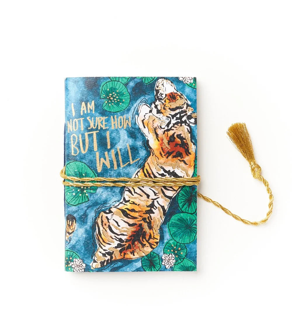 A photo of s journal that is made from recycled cotton in India. This one has a cover with a tiger in water with lily-pads, and the quote "I am not sure how but I will" with a gold colored tassel wrapped around the journal. 