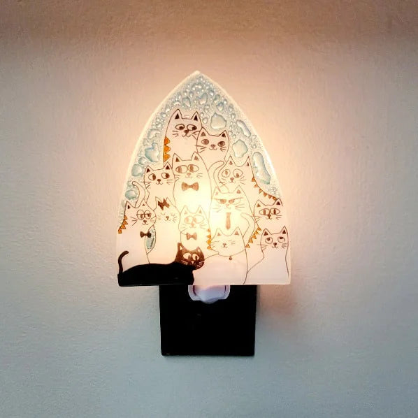 A photo of a nightlight made of recycled glass in Ecuador. This night light features a ton of black and white kitties with ties and different expressive features. 