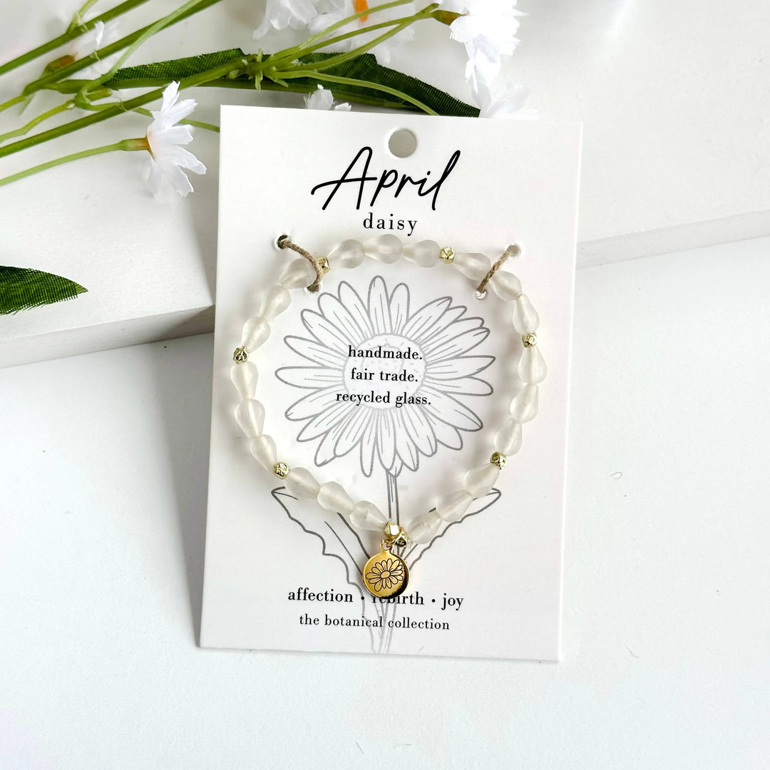 A photo of a white bracelet made from recycled glass in India with a botanical gold-plated charm symbolizing Daisy, the flower of that birth month, April,  on a colored card with the meaning of that flower. 