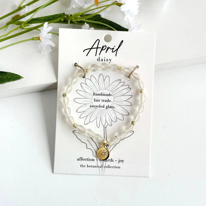 A photo of a white bracelet made from recycled glass in India with a botanical gold-plated charm symbolizing Daisy, the flower of that birth month, April,  on a colored card with the meaning of that flower. 