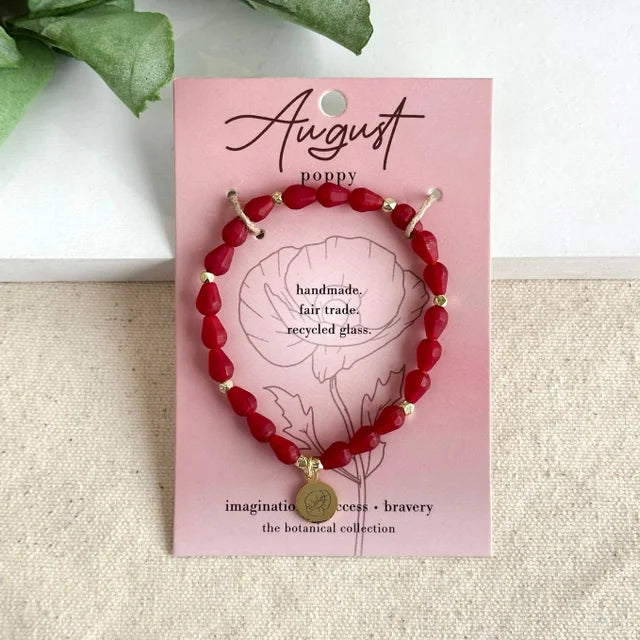 A photo of a red bracelet made from recycled glass in India with a botanical charm symbolizing Poppy, the flower of that birth month, August, on a colored card with the meaning of that flower. 