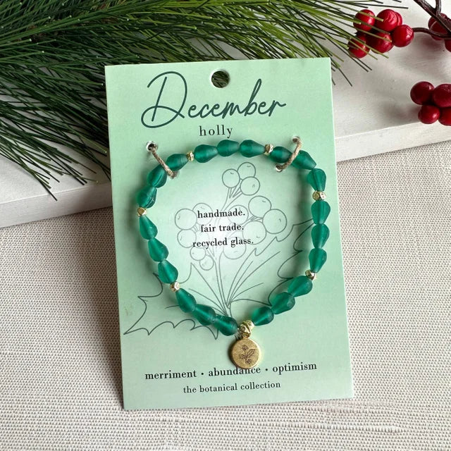 A photo of a green bracelet made from recycled glass in India with a botanical charm symbolizing Holly, the flower of that birth month, December, on a colored card with the meaning of that flower. 