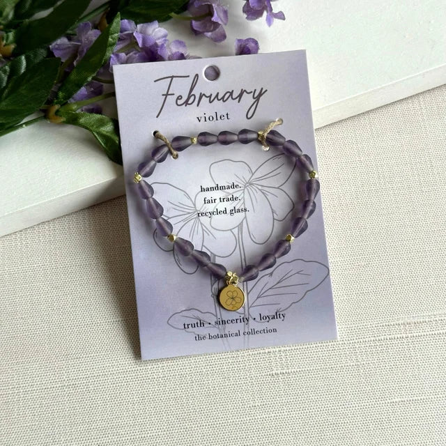 A photo of a purple bracelet made from recycled glass in India with a botanical charm symbolizing Violet, the flower of that birth month, February, on a colored card with the meaning of that flower. 