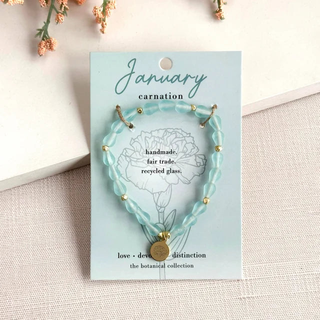 A photo of a light blue bracelet made from recycled glass in India with a botanical charm symbolizing Carnation, the flower of that birth month, January, on a colored card with the meaning of that flower. 
