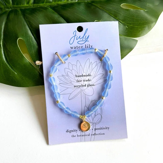 A photo of a blue bracelet made from recycled glass in India with a botanical gold-plated charm symbolizing Water Lily, the flower of that birth month, July, on a colored card with the meaning of that flower. 