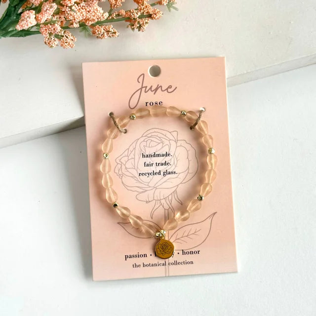 A photo of a blush-colored bracelet made from recycled glass in India with a botanical gold-plated charm symbolizing Rose, the flower of that birth month, June, on a colored card with the meaning of that flower. 
