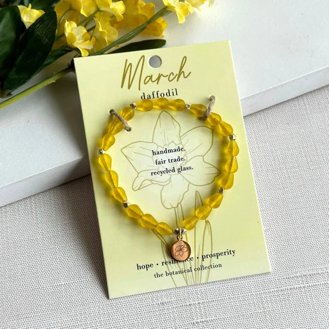 A photo of a yellow bracelet made from recycled glass in India with a botanical gold-plated charm symbolizing Daffodil, the flower of that birth month, March, on a colored card with the meaning of that flower. 