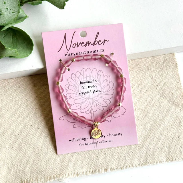 A photo of a pink bracelet made from recycled glass in India with a botanical gold-plated charm symbolizing Chrysanthemum, the flower of that birth month, November, on a colored card with the meaning of that flower. 