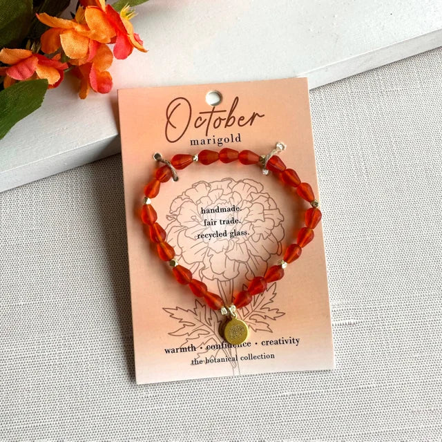 A photo of a orange bracelet made from recycled glass in India with a botanical gold-plated charm symbolizing Marigold, the flower of that birth month, October,  on a colored card with the meaning of that flower. 