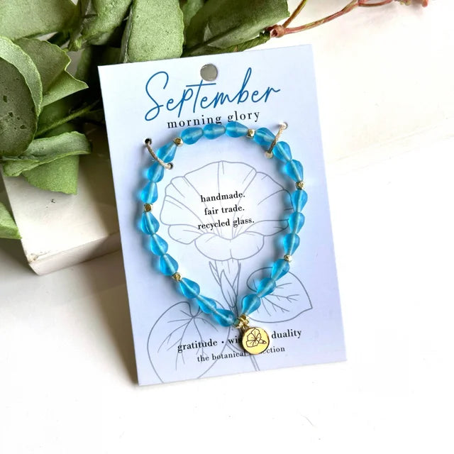 A photo of a blue bracelet made from recycled glass in India with a botanical gold-plated charm symbolizing Morning Glory, the flower of that birth month, September, on a colored card with the meaning of that flower. 