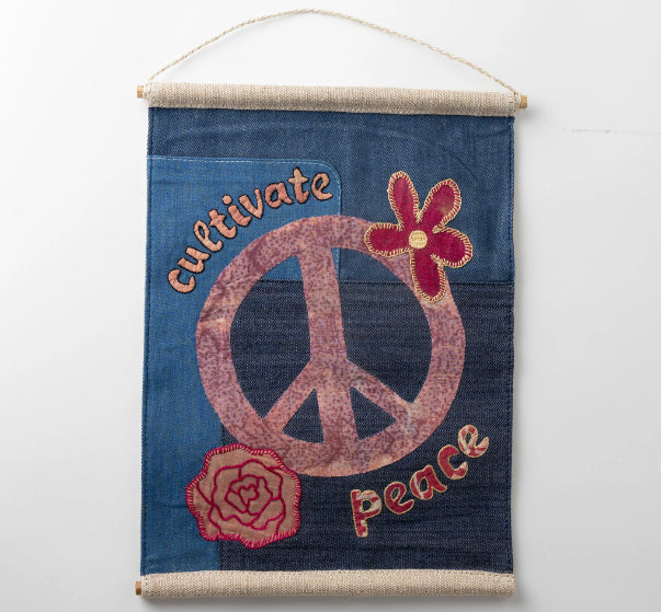 A photo of a wall hanging that reads 'Cultivate Peace' with a Peace Symbol and flowers decorating it. The background is a denim fabric and all the design is made from colorful recycled sari material. 