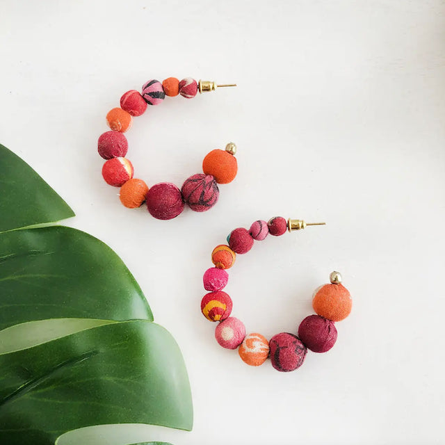 A photo of a pair of earrings that are hoops with circle beads that grow larger as the hoop continues. Each bead is covered in pink/red/orange colored sari material. 