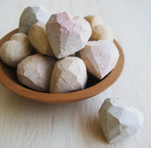 A photo of a bowl with rough edge hearts in it with one heart sitting on the table. All are natural colors of the soapstone properties and in progress of being finished into hearts. 