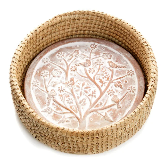 A photo of a bread warmer that is a terra cotta stone in a circle shape with a white washed Birds of a Feather design with a tree and birds and flowers on it, in a kaisa grass basket. 