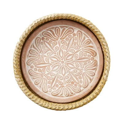 A photo of a bread warmer that is a terra cotta stone in a circle shape with a white washed mandala lotus design on it, in a kaisa grass basket. 