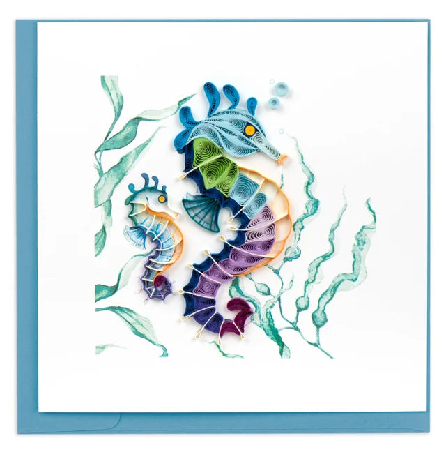 A photo of a greeting card designed with quilling art, the art of rolled paper in Vietnam, on the cover. This card showcases two seahorses, one large seahorse in the foreground with colorful features of varying shades of blue, green, purple, and orange,  with a smaller seahorse next to it and seaweed watercolor in the background of the card. 