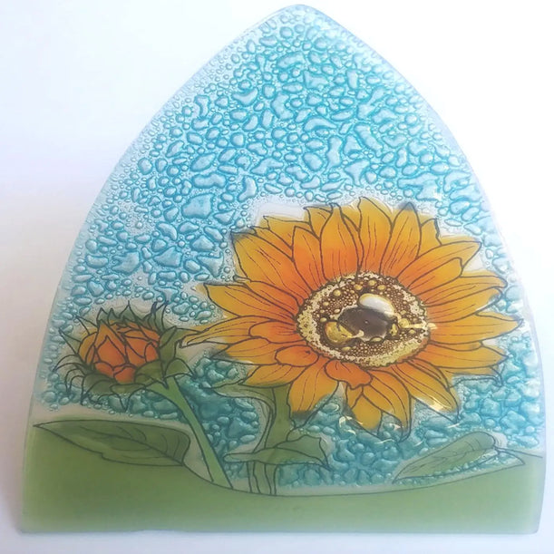 A photo of a nightlight made of recycled glass in Ecuador. This night light features a open sunflower in the forefront, and a closed about to bloom sunflower behind it with a blue and green background. 