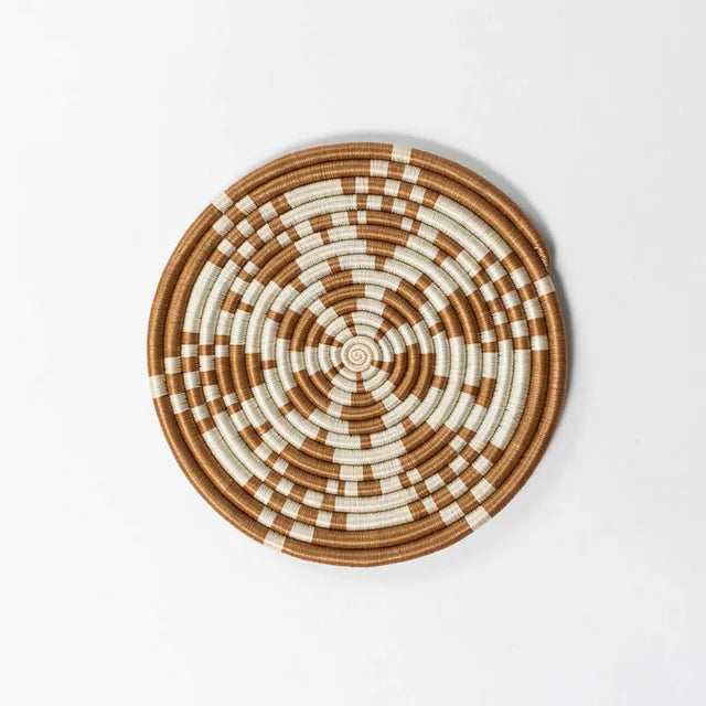 A photo of a trivet made from grass in Rwanda. This one is a tea color with a cream white in a kaleidoscope design. 