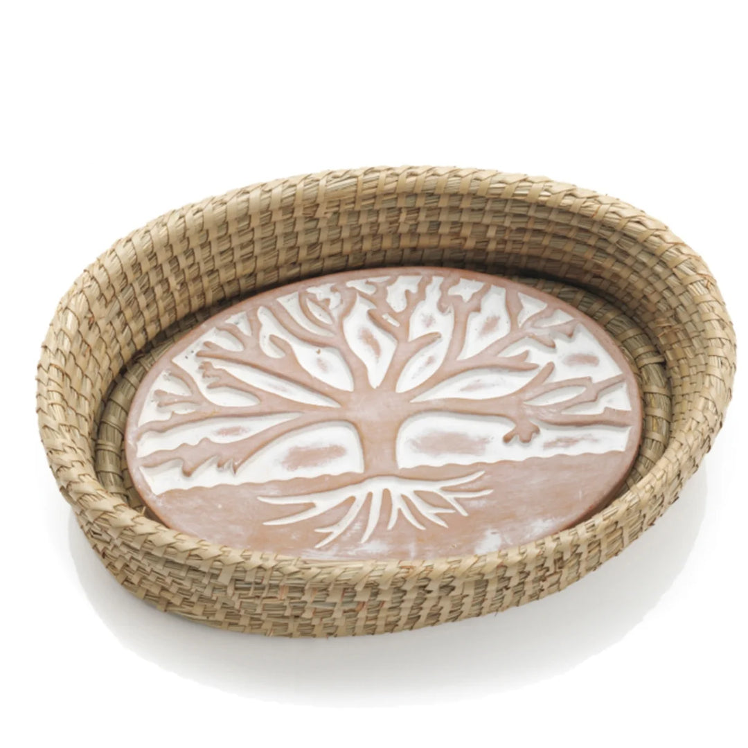 A photo of a bread warmer that is a terra cotta stone in a oval shape with a white washed tree of life on it, in a kaisa grass basket. 